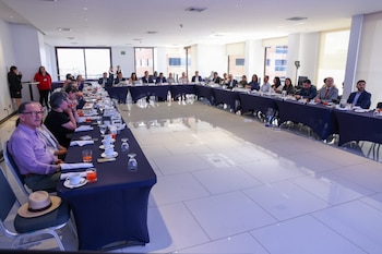 Vista interior de una sala de conferencias luminosa con un grupo de personas sentadas alrededor de una mesa grande en forma de U, con bebidas y papeles