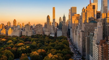 Vista aérea do Central Park com suas folhas de outono e ruas movimentadas, cercado por arranha-céus iluminados pela luz dourada de um pôr do sol nova-iorquino.