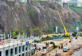 Un acantilado rocoso, una grúa amarilla de construcción, edificaciones en obra, una carretera, palmeras, vehículos y personas