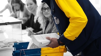 Un trabajador postal con uniforme amarillo y azul sostiene un paquete de documentos sobre una caja cerca de una urna de votación.