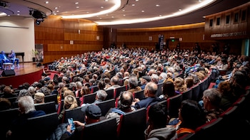 Un hombre habla con un micrófono sobre el escenario frente a un público numeroso que ocupa las butacas de un auditorio de madera