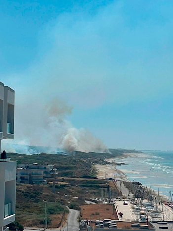 Vista aérea de una zona costera con edificios, dunas, playa y mar, donde columnas de humo se elevan desde la vegetación junto a un puerto con barcos