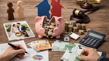 Una mesa de madera con una mano izquierda escribiendo, billetes de euro, un cerdito roto con monedas, una foto de boda, un mazo y figuras de madera.