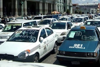 Numerosos taxis blancos y azules estacionados en una calle de ciudad. Algunos exhiben mensajes escritos en sus parabrisas. Se ven edificios y personas