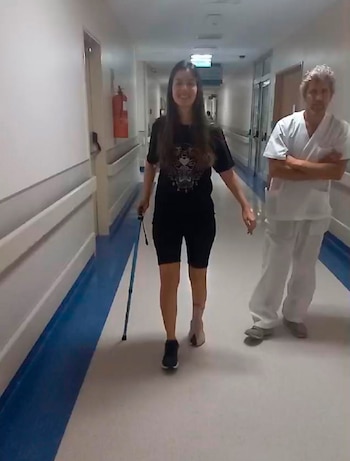 Manuela Lori sonríe caminando con bastón por pasillo de hospital. Viste oscuro, un zapato y vendaje en un pie. Un hombre de uniforme médico la acompaña