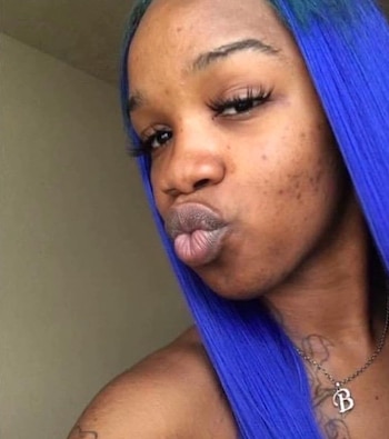 Retrato de una mujer con el cabello teñido de azul, una cadena con la letra B y la boca en postura de beso