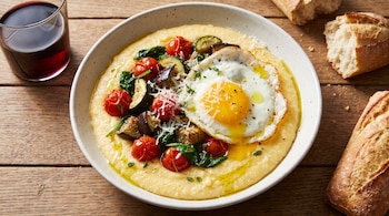 Plato de polenta con huevo frito, verduras (tomates, calabacín, berenjena, espinaca), queso y tomillo, sobre mesa de madera. Una copa de vino tinto y pan.