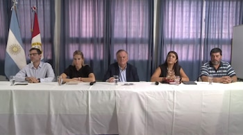 Cinco pessoas, três homens e duas mulheres, sentam-se atrás de uma mesa branca com um copo de água e um microfone. Duas bandeiras estão ao fundo, perto da cortina roxa