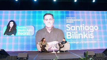 María Julia Bearzi y Santiago Bilinkis sentados en sillones grises en un escenario con una pantalla gigante de fondo que muestra sus nombres