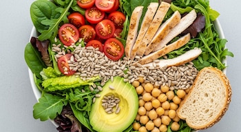 Vista cenital de un bowl blanco lleno de ensalada fresca con pollo grillado, tomates cherry, hojas verdes, palta, garbanzos, semillas de girasol y pan integral.