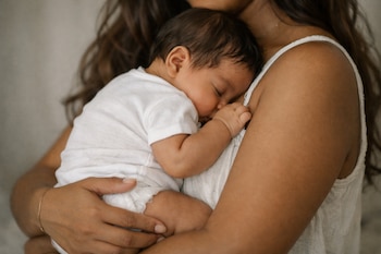 Primer plano de una madre latina sosteniendo a su bebé durmiendo en su pecho, ambos vestidos de blanco. Se aprecian sus brazos y el torso de la madre.