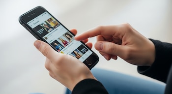 Primer plano de dos manos sosteniendo un smartphone negro, con un dedo índice tocando la pantalla que muestra un feed con varias imágenes de personas.