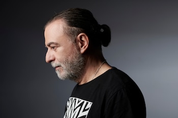 Un hombre con barba gris y cabello oscuro recogido en un moño se muestra de perfil, vistiendo una camiseta negra con diseño y una cadena plateada sobre un fondo gris liso