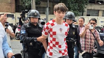 Un hombre joven con camiseta a cuadros rojos y blancos es escoltado por dos policías de uniforme oscuro. Se observan personas con teléfonos, micrófonos y motocicletas