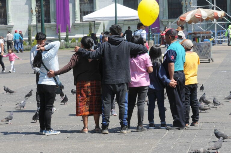 Una familia, incluyendo dos adultos y una niña pequeña, camina de espaldas por una acera concurrida, con comercios que muestran nombres en inglés y una calle adyacente.