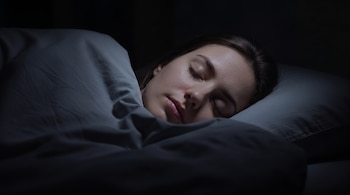 Mujer joven durmiendo en una cama; cubierta por una manta gris; cabeza apoyada en almohada; habitación oscura.