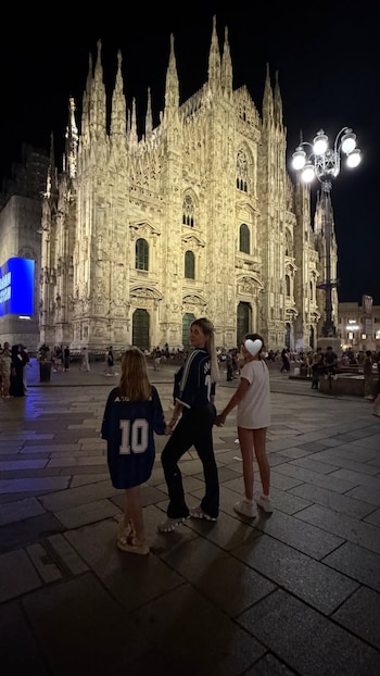 Tres mujeres de espaldas, una de ellas con cabello rubio, frente a la fachada iluminada de una catedral gótica por la noche en una plaza pavimentada con personas
