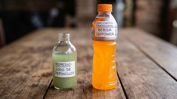 Dos botellas en una mesa de madera: una pequeña con líquido verde y etiqueta "Remedio: Jugo de Pepinillos", y una grande de bebida isotónica naranja.