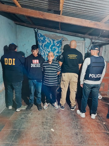 Cinco hombres de pie en un interior con paredes azules y techo de chapa. Dos oficiales de policía y tres individuos detenidos, dos con caras pixeladas