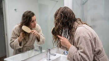 Mujer con bata beige se cepilla el cabello ondulado, mojado y castaño, frente a un espejo empañado con gotas de agua; se ve el reflejo y un grifo.