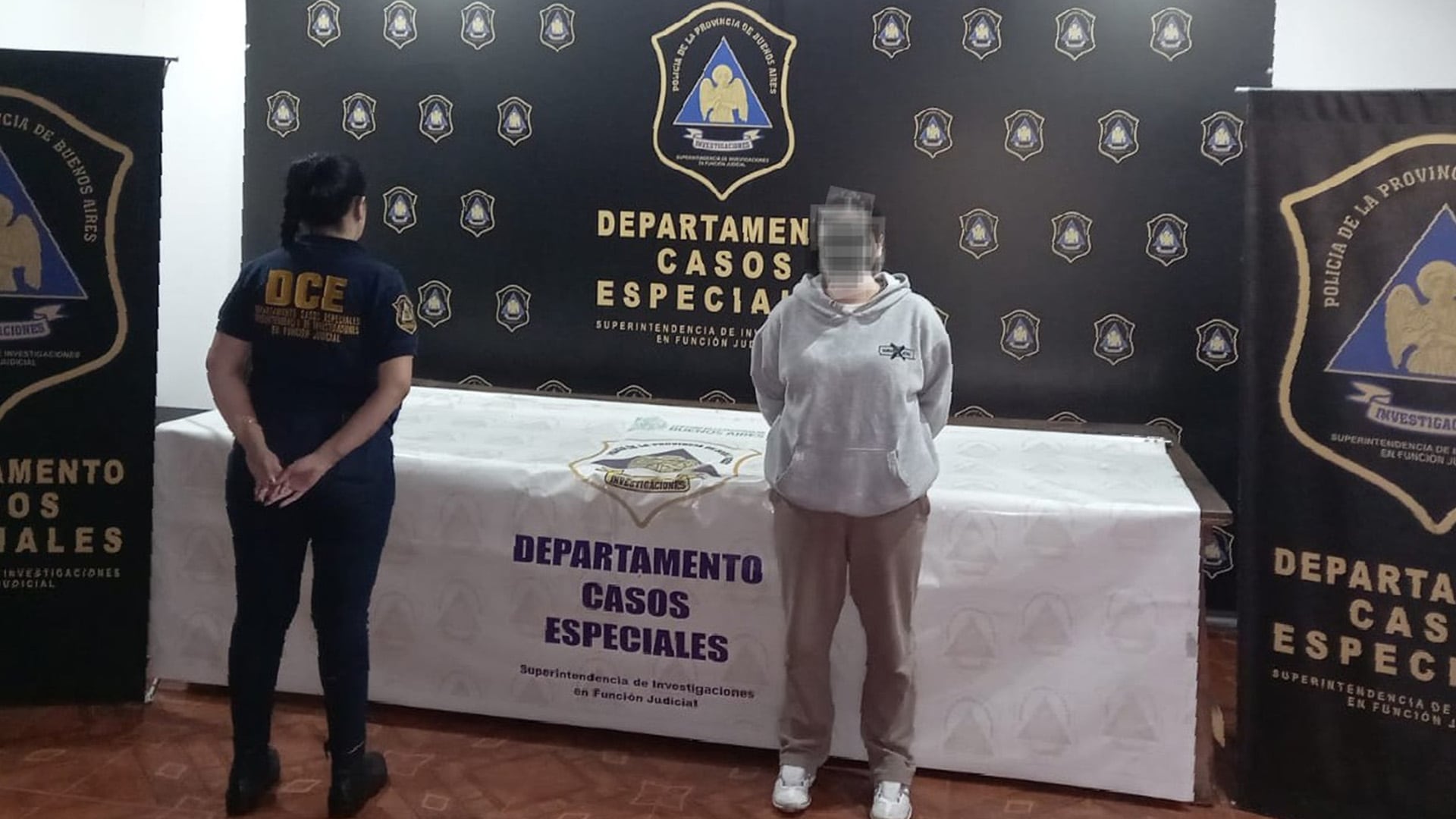 Una oficial del Departamento de Casos Especiales de la Policía de la Provincia de Buenos Aires y una persona detenida posan frente a un banner identificatorio del organismo en una presentación oficial.
