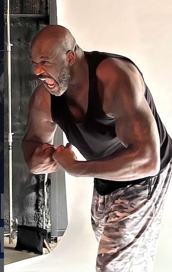 Terry Crews, un hombre calvo y barbudo, viste una camiseta negra sin mangas y pantalones cortos de camuflaje, flexiona sus bíceps y grita