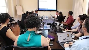 Un grupo de personas sentadas alrededor de una mesa de reuniones, usando laptops y observando una gran pantalla con gráficos al fondo