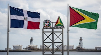 Banderas de República Dominicana y Guyana ondeando flanqueando logos nacionales. Al fondo, instalaciones petroleras con torres y tanques de extracción sobre el mar.