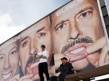 Valla publicitaria con imágenes de tres rostros masculinos y textos en los labios. Dos hombres posan sobre la estructura. Al fondo, carretera con coches y montañas