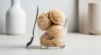 Cuatro bolas de helado de café y leche de almendras dentro de un vaso transparente con una cuchara plateada a un lado. Fondo blanco y minimalista con botellas desenfocadas.