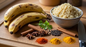 Primer plano de plátanos machos, un cuenco de quinoa cocida y pilas de especias (canela, anís, pimienta, pimentón, cúrcuma) sobre tabla de madera.