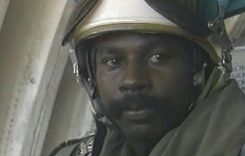 Un hombre de piel oscura con bigote y un casco de vuelo blanco y verde con visera levantada mira al frente, mostrando su rostro en primer plano