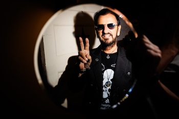 Ringo Starr lleva gafas de sol y chaqueta negra mientras hace el signo de la paz con la mano, visto a través de un marco circular