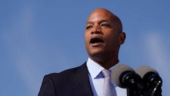 Primer plano de Wes Moore, un hombre calvo de piel oscura, hablando frente a dos micrófonos con un traje oscuro y corbata estampada bajo un cielo azul claro