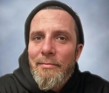 Un hombre caucásico de mediana edad con gorro oscuro, barba y bigote mira al frente; el fondo es un suave degradado azul y gris