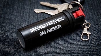 Un espray negro de gas pimienta con botón rojo de seguridad, conectado a un llavero con dos llaves, sobre tela oscura.