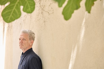 Un hombre de cabello canoso y camisa azul oscuro mira a la izquierda, apoyado en una pared beige texturizada, con hojas verdes en la parte superior