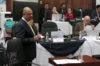 Un hombre calvo vestido con traje oscuro y corbata amarilla habla frente a un micrófono en un salón con otras personas y laptops sobre mesas