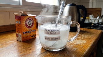 Caja de bicarbonato de sodio junto a una taza de vidrio con bicarbonato y agua caliente burbujeando, una cuchara dentro y una tetera eléctrica al fondo en una cocina.