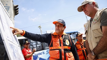 Dos hombres con chalecos y gorras observan un mapa sobre un caballete en la calle. Uno señala el mapa. Hay un autobús y otras personas al fondo