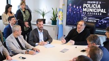 Hombres y mujeres en sala de reuniones alrededor de una mesa. Un hombre gesticula al hablar, con una pantalla al fondo que muestra información sobre la Policía de Escobar