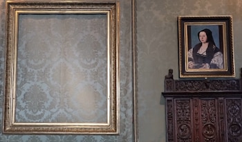 Un gran marco dorado y vacío cuelga a la izquierda de una pared con papel tapiz. A la derecha, un retrato enmarcado de una mujer sobre un mueble tallado de madera oscura
