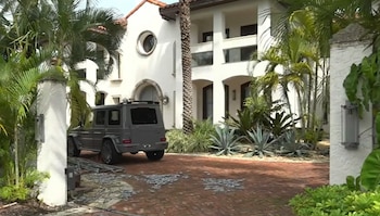 Fachada de una casa blanca de dos pisos con un vehículo todoterreno gris estacionado en una entrada de ladrillos rojos rodeada de palmeras