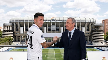 Enzo Fernández con camiseta blanca número 24 y Florentino Pérez de traje, estrechando manos frente a micrófonos, con el estadio Santiago Bernabéu de fondo.
