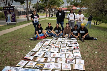 Grupo de manifestantes, mayormente mujeres, se sienta y de pie sobre césped junto a decenas de fotos de individuos y pancartas que exigen libertad, con árboles