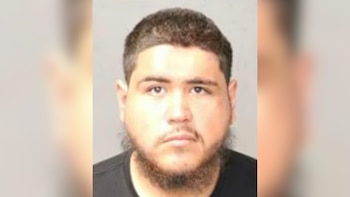 Fotografía policial de un hombre joven con cabello corto oscuro, barba en la barbilla y camiseta negra frente a un fondo neutro