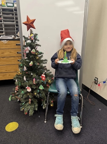 Una niña sentada en una silla sostiene un plato blanco junto a un árbol de Navidad decorado con luces y una estrella