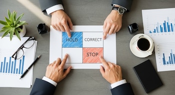 Vista cenital de dos manos sobre una mesa señalando una matriz con los términos Hold, Correct y Stop. Incluye una planta, anteojos, bolígrafo, gráficos y una taza de café.