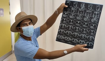 Hombre con sombrero y mascarilla sostiene una placa grande con imágenes médicas. Tiene vendajes en ambos brazos. Fondo claro, puerta amarilla con cartel de baño