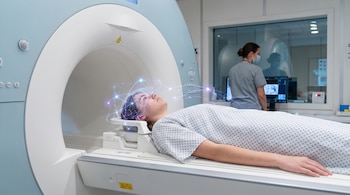 Un joven recostado en una camilla de resonancia magnética con luces sobre la cabeza y una profesional médica frente a monitores al fondo.